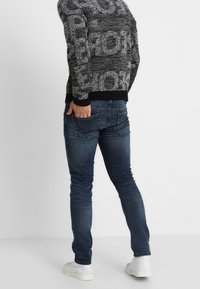 Jeans slim-fit bleu foncé avec un effet délavé, dotés de deux poches arrière et de coutures subtiles, associés à un pull à motifs noir et gris.