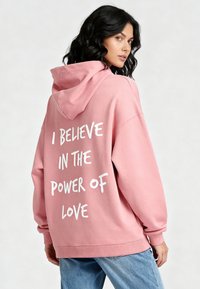 Felpa con cappuccio rosa in cotone con una grande scritta bianca stampata sulla schiena, dotata di cappuccio con coulisse e vestibilità ampia. Abbinata a jeans azzurri.