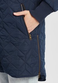 Tengerész navy színű steppelt kabát, gyémánt mintázattal, bordázott mandzsettákkal és arany cipzár részletekkel; oldalán látható zseb, amibe egy kéz van betéve.