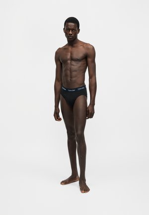 Άνδρας μοντέλο με σκούρο δέρμα και κοντά μαλλιά, φορώντας μαύρα Calvin Klein εσώρουχα, ποζάρει μπροστά σε απλό φωτεινό φόντο.