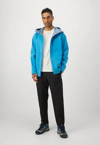 Giacca impermeabile blu con interno grigio chiaro, cerniera frontale, abbinata a pantaloni neri e scarpe da ginnastica blu, indossata da una figura in piedi.