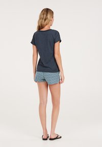 Maglietta a maniche corte blu navy con maniche arrotolate, abbinata a pantaloni shorts turchesi a fantasia, caratterizzati da un design geometrico e una vestibilità aderente. Infradito nere.