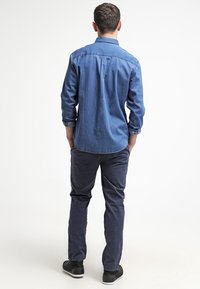 Camisa de mezclilla azul, de manga larga, con cuello abotonado y detalle de pliegue en la parte posterior. Combinada con pantalones oscuros navy y zapatillas negras.