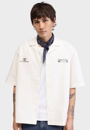 Young person with short brown hair wearing a white short-sleeve button shirt, a blue patterned neck scarf, and visible arm tattoos.