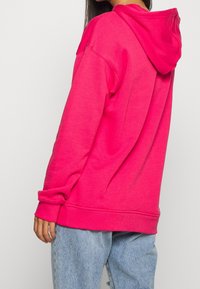 Rosa bomullshoodie med dragsko i huven, sänkta axlar, ribbade muddar och avslappnad passform, kombinerad med ljusblå jeans.