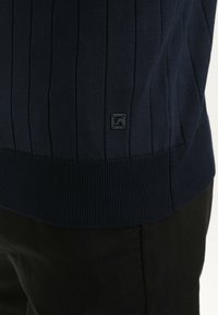 Marineblauwe geribbelde trui met een gestructureerd breipatroon en een klein zwart logo-label near de zoom, gecombineerd met zwarte broek.