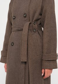 Brun trenchcoat i høringsbenmønster med bælte, dobbeltknap design, sorte knapper og manchetter med opsmøg lavet af tekstureret stof.