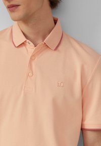 Pfirsichfarbendes Poloshirt aus strukturiertem Stoff, mit einem Kragen in Pinktönen, drei Knöpfen und einem dezenten Logo auf der linken Brust.