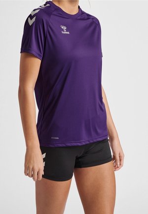 Kobieta ubrana w fioletową sportową koszulkę hummel i czarne spodenki, stojąca z rękami swobodnie opuszczonymi wzdłuż ciała na jednolitym tle.