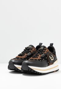 Sneaker nero e marrone che presenta una combinazione di pelle e camoscio, con dettagli dorati, superficie testurizzata e una suola a strisce bianche e dorate.