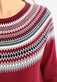 Pull en maille rouge avec motif géométrique en zigzag et diamants blanc, noir et rose sur la poitrine et col rond.