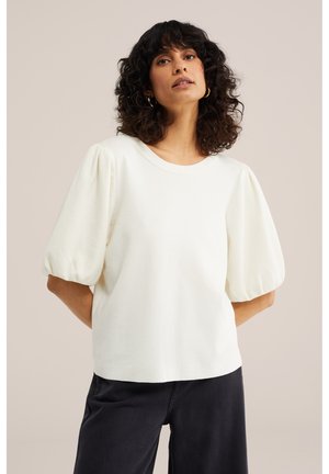 Mujer con el pelo rizado que lleva pendientes de aro dorados, una blusa blanca con mangas abullonadas y pantalones negros, de pie frente a un fondo liso.