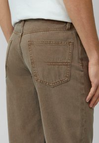 Los jeans de mezclilla marrones presentan un corte recto, bolsillos traseros con costuras sutiles y una ligera textura, combinados con una camisa de color claro.