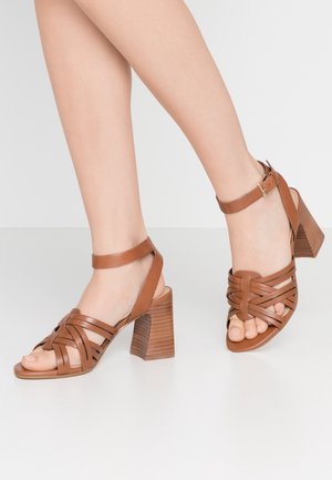 Brown leather heeled sandals with a woven strap design, open toe, ankle strap, and ribbed block heel. Features a smooth, polished texture.