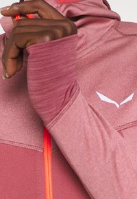 Rosa träningsjacka med en strukturerad tyg, kontrasterande orange dragkedja och en vit logotyp på bröstet. Tummhål i ärmarna.