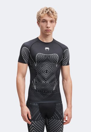 Jeune homme portant un t-shirt de compression noir et des leggings avec des motifs géométriques argentés, debout devant un fond clair uni.