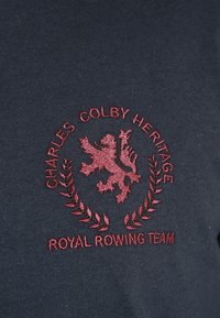 Dark navy fabric featuring red embroidered crest with a lion and laurel leaves, text reads "CHARLES COLBY HERITAGE" and "ROYAL ROWING TEAM."