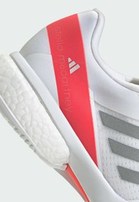 White athletic shoe featuring a textured white sole, gray stripes, and a red accent area with "stella mccartney" branding.