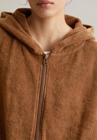 Veste à capuche brune à zip fabriquée en tissu doux et texturé. Dotée d'une fermeture éclair à l'avant, de coutures visibles et d'une coupe décontractée.