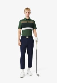 Man som har på sig en randig polo i grönt, vitt och oliv, marinblå byxor, vita skor och en golfhandske, håller en golfklubba framför en vit bakgrund.