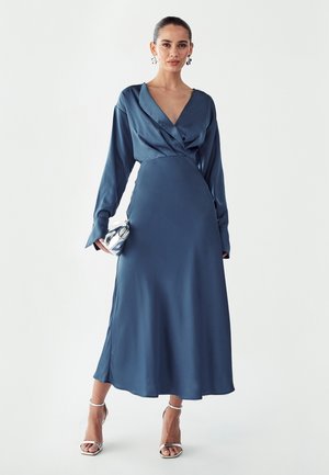 Šaty s dlhým rukávom a zavinovacím strihom z hlboko modrého saténu, s výstrihom do V a splývavou siluetou. Kombinované so striebornými sandálmi na tenkých remienkoch a kabelkou do ruky.