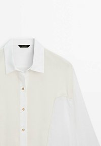 Chemise blanche à manches longues avec un gilet beige en tricot par-dessus et des boutons dorés sur le devant, présentée sur un fond uni.
