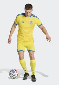 Completo da calcio giallo con motivi geometrici, accenti blu sul colletto e sulle maniche, abbinato a scarpe da calcio nere e una palla multicolore.