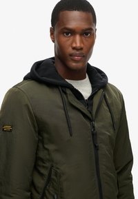 Giacca leggera verde oliva con cappuccio nero, chiusura con zip, coulisse e patch Superdry sulla manica sinistra.