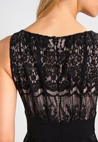 Robe en dentelle noire avec des motifs floraux, dos transparent, et une taille noire unie. Texture détaillée et design élégant. Fermeture éclair dans le dos.