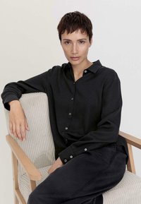 Schwarze Hemdjacke mit weicher Textur und langen Ärmeln, kombiniert mit passenden dunklen Hosen, sitzt auf einem hellen, gerippten Stoffstuhl.