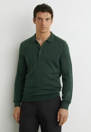 Man draagt een donkergroene poloshirt met lange mouwen met verticale strepen en een zwarte broek, staand tegen een witte muur.
