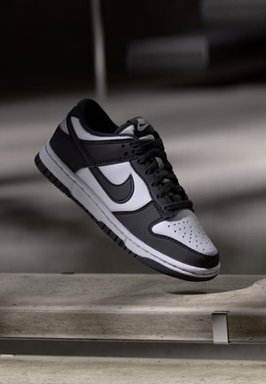Sort-hvid Nike-sneaker med snørebånd, der svæver over en betonoverflade mod en sløret mørk baggrund.