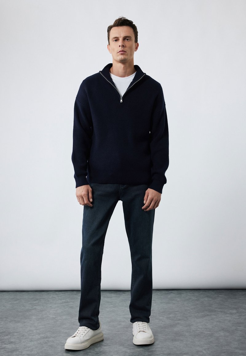 Suéter de punto en azul marino con cuello con cremallera, combinado con jeans de mezclilla oscura y zapatillas blancas, con un ajuste relajado y textura acanalada.