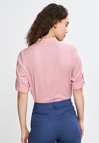 Różowa bluzka z kołnierzykiem i podwiniętymi rękawami, wykonana z gładkiego materiału. Połączona z niebieskimi spodniami o wysokim stanie z tylnymi kieszeniami.