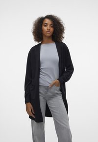 Zwarte open voor cardigan met lange mouwen, twee voorzakken, gedragen boven een lichtgrijs top. Het model draagt ook lichtgrijze wijde broekspijpen.