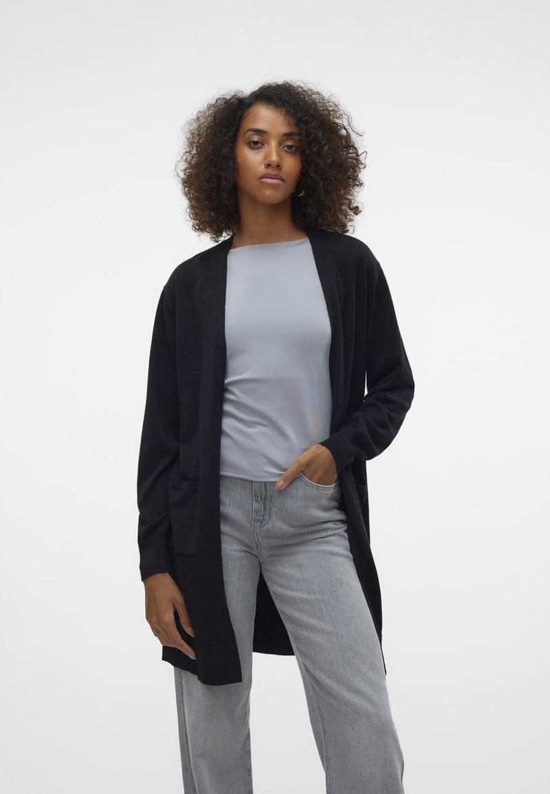 Zwarte open voor cardigan met lange mouwen, twee voorzakken, gedragen boven een lichtgrijs top. Het model draagt ook lichtgrijze wijde broekspijpen.