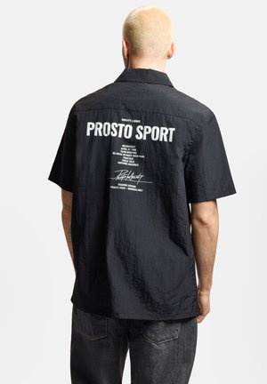 Osoba z krótkimi blond włosami w czarnej koszulce z krótkim rękawem z białym napisem "PROSTO SPORT" i grafiką na plecach.
