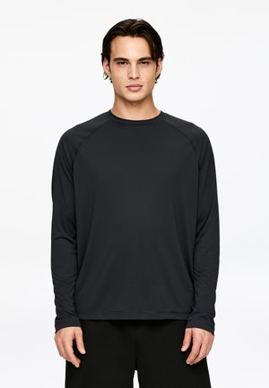 Young man with dark hair wearing a plain long-sleeve black shirt and black pants, standing against a white background, facing forward.