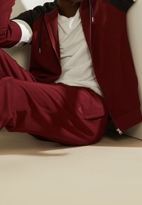 Chándal granate con chaqueta con cremallera que presenta detalles en negro en los hombros, combinado con pantalones a juego. Textura suave y diseño estilizado.