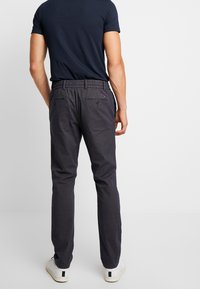 Muž v tmavě modrých slim fit kalhotách a černém tričku, stojící s rukama podél těla, čelící hladkému bílému pozadí.