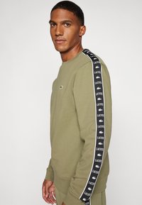 Sudadera verde oliva con cuello redondo, que presenta una cinta de marca en blanco y negro a lo largo de las mangas y un pequeño logo en el pecho.