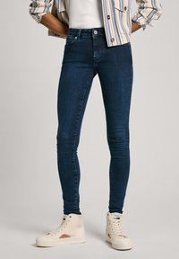 Jeans ajustados de color azul oscuro hechos de denim, con un bolsillo frontal, detalles de costura sutiles y combinados con zapatillas altas de color claro.