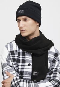 Zwarte gebreide muts en bijpassende sjaal gedragen bij een zwart-witte geruite shirt. Label op de sjaal leest "I SOLID." Zachte texturen, eenvoudig ontwerp.