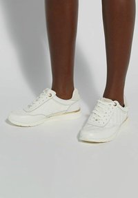 White sneakers with quilted texture, gold accents, and a cushioned sole. Laces and rounded toe add practical detail.