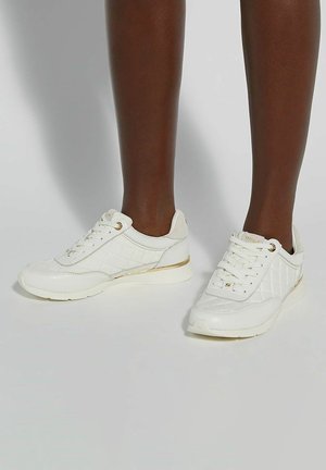 White sneakers with quilted texture, gold accents, and a cushioned sole. Laces and rounded toe add practical detail.