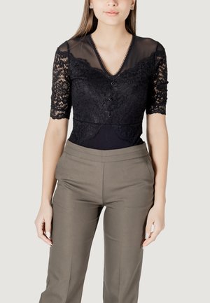 Blusa nera in pizzo con maniche corte trasparenti e scollo a V, caratterizzata da bottoni decorativi e dettagli in pizzo. Abbinata a pantaloni beige aderenti.