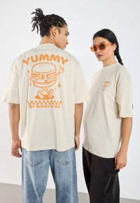 Beige oversized t-shirts with orange graphics featuring a hamburger and text, paired with relaxed jeans and sunglasses.