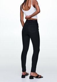 Jeans negros ajustados con cintura alta, con bolsillos traseros estándar y una textura suave. Combinados con simples sandalias negras de tipo deslizante.
