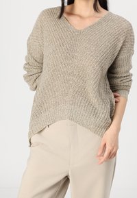 Femme portant un pull en maille beige texturé avec un col en V et un pantalon beige clair, posant avec une main sur la hanche.