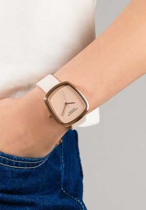 Poignet portant une montre à cadran carré avec un cadran beige, un boîtier en or rose et un bracelet blanc, main partiellement dans la poche d'un jean bleu.
